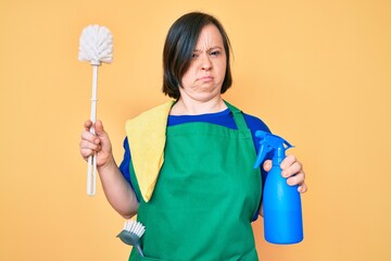 Brunette woman with down syndrome wearing apron holding scourer and toilet brush skeptic and nervous, frowning upset because of problem. negative person.