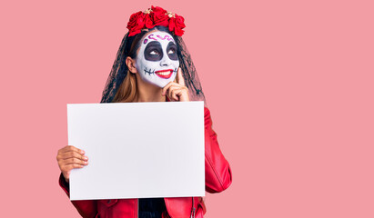 Woman wearing day of the dead costume holding blank empty banner serious face thinking about question with hand on chin, thoughtful about confusing idea