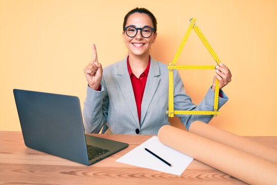 Young brunette architect woman at the office holding build project smiling with an idea or question pointing finger with happy face, number one