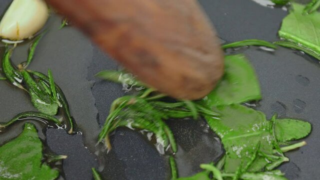 Close-up frying garlic cloves, rosemary and sage in oil into a frying pan. Concept of cooking healthy by preparing greens in 4K.