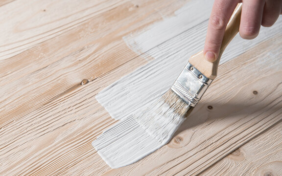 Man hand paints wooden surface with brush and white paint. Natural wooden texture. Renovation or repair construction. 