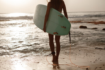 Cropped image of active woman dressed in swimsuit, carries surf board, walks on coastline against sunshine, ready to catch waves. Surfing, water sport and recreation concept