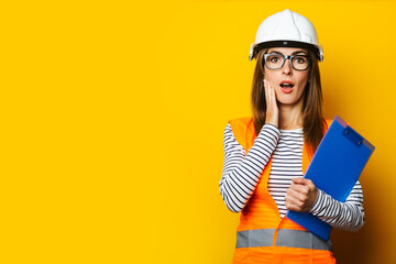 Young woman with a surprised face in a vest and hard hat holds a clipboard on a yellow background. Construction concept, new building. Banner