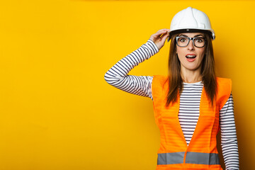Young woman with a surprised face in a vest and hard hat on a yellow background. Construction...