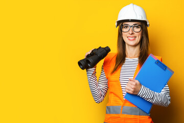 Young woman with a smile in a vest and hard hat holds a clipboard and binoculars on a yellow background