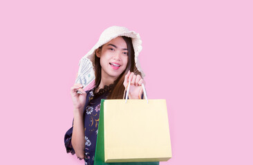 Fototapeta premium Asian young woman with a lot of money in her hand holding a shopping bag smiling happily on the pink background.