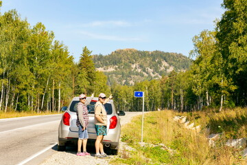a man and a woman autotourists stand on the highway near the car and point to the high mountain...
