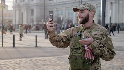 Confident soldier with a beard and american flag having video call with family using mobile phone after war zone or training outdoors. - Powered by Adobe