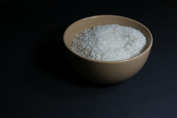 White uncooked rice in a bowl with scattered grains nearby on a black background