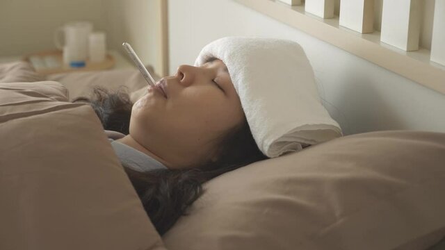 Side view, panning shot of sick Asian woman Lying on bed, with towel on forehead to reduce relieve fever and reduce body temperature, with oral thermometer in her mouth. Covid-19 concept