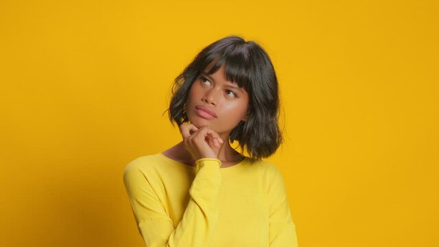 Thoughtful young mixed race woman looks somewhere pensively thinks deeply about something tries to make decision in mind wears casual jumper poses against gold yellow background. Let me think