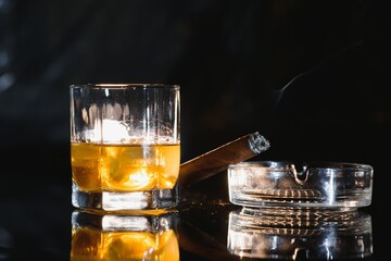 A glass of whiskey or Bourbon with ice cubes and a cigar on a black slate with smoke