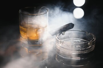 Glass of whiskey with ice cubes and smoking cigar on table