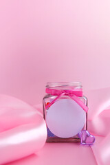 Glass jar with candy hearts inside and a pink silk ribbon on a pink background. Concept for Valentine's Day. Mockup with a place for a logo. Vertical banner. Side view. Copyspace