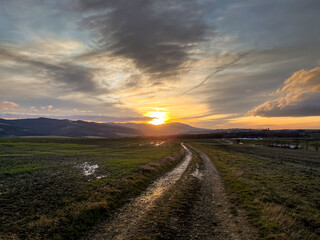 A dirt road leading around a field during an orange sunset on the horizon of the setting sun behind the hills.