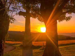 The setting sun over the landscape overlooking the trees and the cross where the sun sets between the objects.