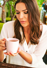 Portrait of beautiful young woman wearing white clothes, relaxing on the balcony between many green plants, holding cup of tea