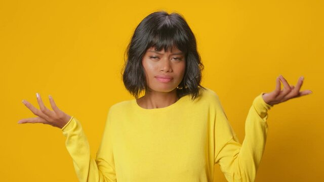 Pretty mixed race woman shrugs shoulders unaware raises hand clueless has no idea cannot answer wears casual jumper poses against gold yellow background. Ethnic beautiful girl not knowing information