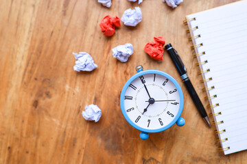 Alarm clock, a pen, a notebook and crumpled papers on the wooden table 