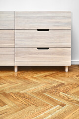 Stylish chest of drawers made with bleached solid oak timber with finger pull sliding stands on parquet floor near white wall