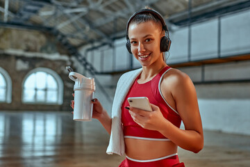 Beautiful woman using smartphone and listening to music in headphones drinks protein shake while...
