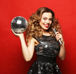 Party, holiday and celebration concept:Young blond woman with long wavy hair dressed in evening dress holding a microphone and disco ball, singing and smiling