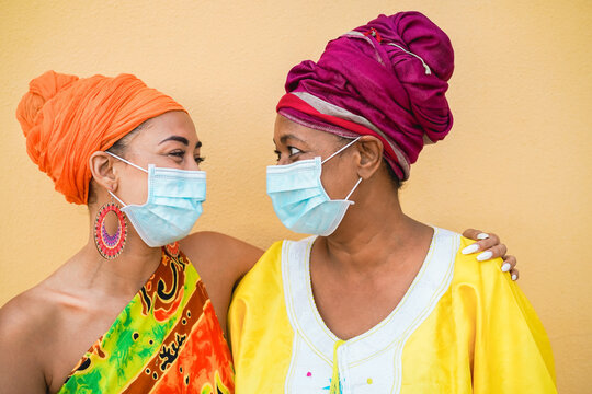 Happy Mother And Daughter Wearing Traditional African Dresses During Coronavirus Outbreak - African People With Face Safety Mask Having Fun - Social Distance Concept - Focus On Faces