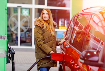 Young caucasian woman at self-service gas station, hold fuel nozzle and refuel the car with petrol, diesel, gas. Pretty woman filling her auto with gasoline or benzine, outdoors. Self service gas pump - obrazy, fototapety, plakaty
