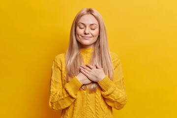 Fototapeta na wymiar Photo of calm blonde Caucasian woman keeps palms pressed to chest expresses gratefulness closes eyes smiles pleasantly has dimples on cheeks being touched by heartwarming words stands indoor