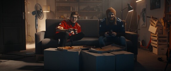 POV Portrait of two friends hanging out, sitting on sofa, playing video game inside garage. Winning...