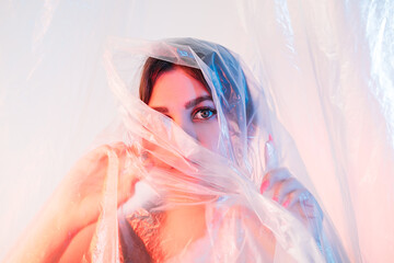 Pandemic self isolation. Quarantine anxiety. Covid-19 lockdown. Stay home. Art portrait of scared curious woman hiding behind transparent wrinkled polyethylene film in iridescent pink blue neon light.