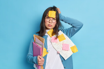 Disappointed forgetful young woman tired of cramming for exam keeps hand on head and looks stressed at camera poses with papers and stickers isolated over blue background has very tired look