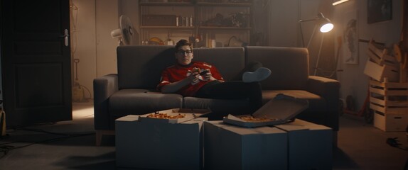 POV Portrait of Caucasian teenager playing video game inside home garage, enjoying pizza. Shot with...