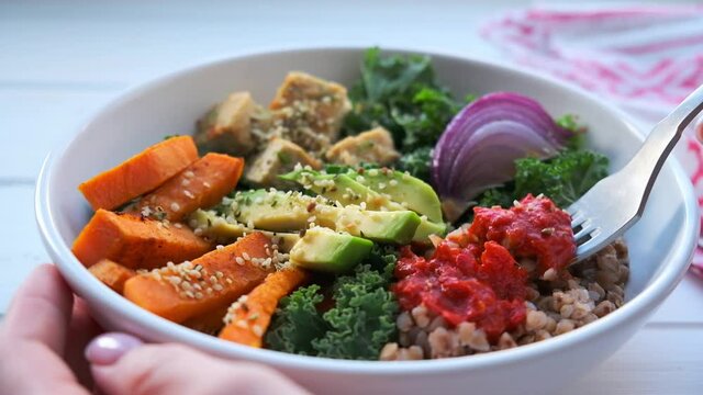 Baked vegetables, avocado, tofu and buckwheat buddha bowl. Vegan lunch salad with kale, baked sweet potato, tofu, buckwheat and avocado in white bowl. Vegan food concept.