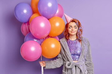 Fototapeta na wymiar Funny birthday girl feels tired after noisy party smiles pleasantly concentrated above smeared with cream has festive mood holds multicolored balloons isolated over purple background. Holidays concept