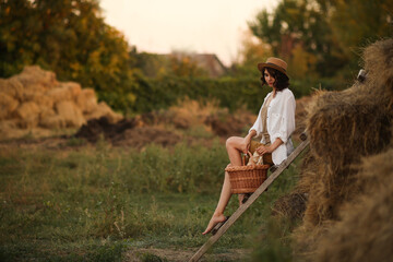 young beautiful girl in a straw hat in a rustic style sits resting in the countryside on an old wooden staircase with a wicker basket in her hand. Vacation relaxation isolation summer