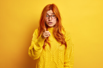 Redhead European woman indicates at you with index finger feels shocked to see something amazing wears transparent glasses and warm sweater isolated over yellow studio background. Omg look ahead