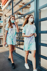 Young women girls spending time together downtown walking along store front in the city center. Girls wearing the face masks to avoid virus infection and to prevent the spread of disease