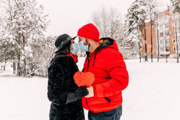Fototapeta na wymiar Young romantic couple in medical masks is having fun outdoors in winter. Snowing. Love concept. Enjoying spending time together. Heart with sequins. Two lovers are hugging in Saint Valentine's 