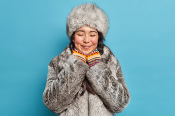 Dreamy satisfied young Asian woman closes eyes and smiles gently wears grey fur hat and coat has...