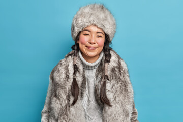 Winter clothes. Pleased Inuit woman with two pigtails smiles gently wears fur hat knitted warm sweater and coat poses against blue background. Happy nenets girl indoor. Aboriginal from north - obrazy, fototapety, plakaty
