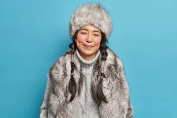 Fotobehang Winter clothes. Pleased Inuit woman with two pigtails smiles gently wears fur hat knitted warm sweater and coat poses against blue background. Happy nenets girl indoor. Aboriginal from north © Wayhome Studio