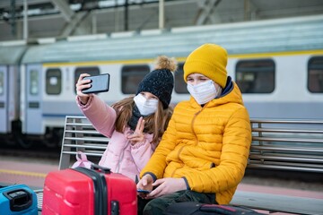 Two children wearing protective masks are sitting on the platform waiting for the train. The girl...
