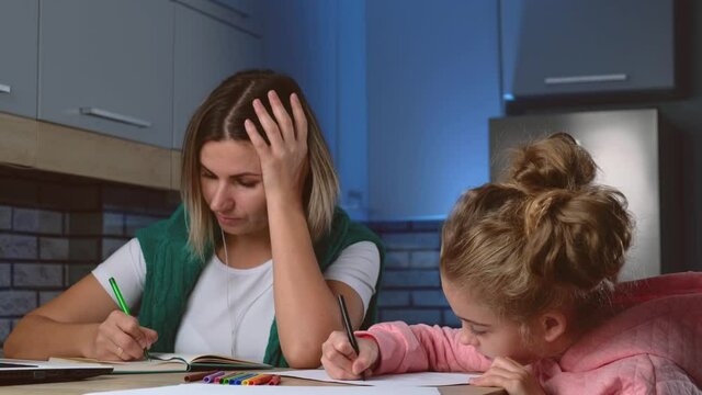 Caucasian mother and daughter are sitting at the table in the living room writing and drawing in copybook with different colors