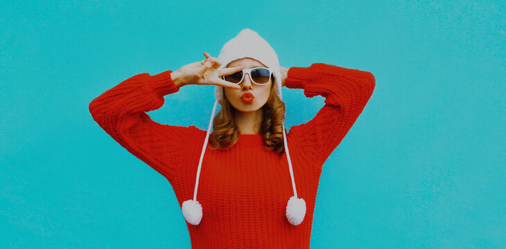 Portrait of young woman blowing red lips wearing a red sweater and hat with pom pom over a blue background