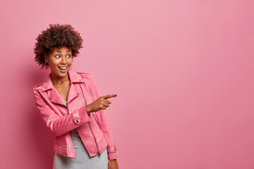 Happy dark skinned curly young woman indicates at distance sees something amazing giggles positively wears stylish jacket isolated over pink background. Wow look at this great offer or sales