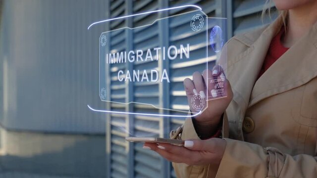 Unrecognizable businesswoman interacts HUD hologram with text Immigration Canada. Woman in the coat uses the technology of the future mobile screen on the background of the city