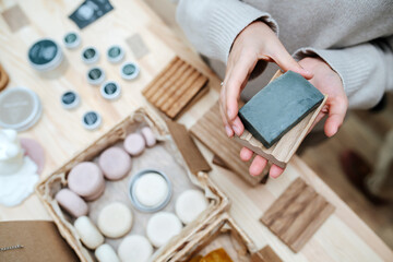 Female hands holding neat eco soap and a wooden soap dish. Overhead view. Standing in ecological shop in front of the table with variety of cosmetic items.