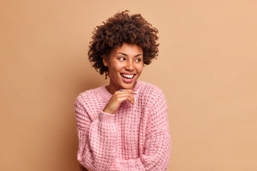 Portrait of curly haired woman keeps hand under chin and looks away gladfully wears knitted jumper has carefree expression poses against brown background. Happy emotions and feelings concept