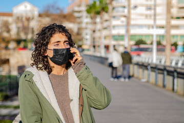 Fototapeta premium Young Lifestyle Man With Worried Expression Face Wearing Black Mask and Green Jacket Talking to Phone in a Bridge at the Street